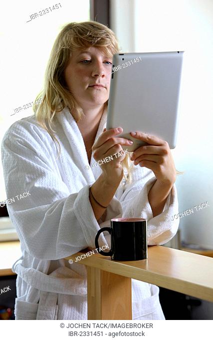 Young woman wearing a dressing gown drinking a cup of coffee in the morning, sitting in the kitchen reading an online newspaper on an iPad, a tablet computer