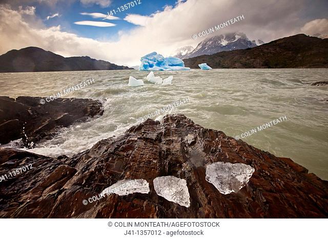 Blue iceberg, rain squalls on Lago Grey, chunks of ice blown onto rocky foreshore, Parque Nacional Torres del Paine, Patagonia, Chile