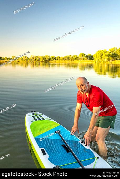 Senior adult paddler is launching inflatable stand up paddleboard on a calm lake in Colorado
