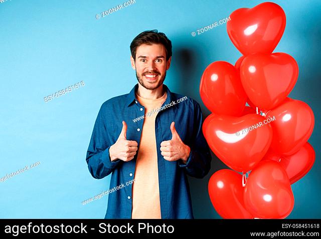 Happy valentines day. Cheerful boyfriend showing thumbs up in approval, standing with red hearts balloons for lover, blue background