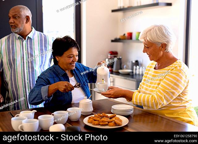 Biracial senior woman serving coffee in cup held by friend at wooden table in nursing home