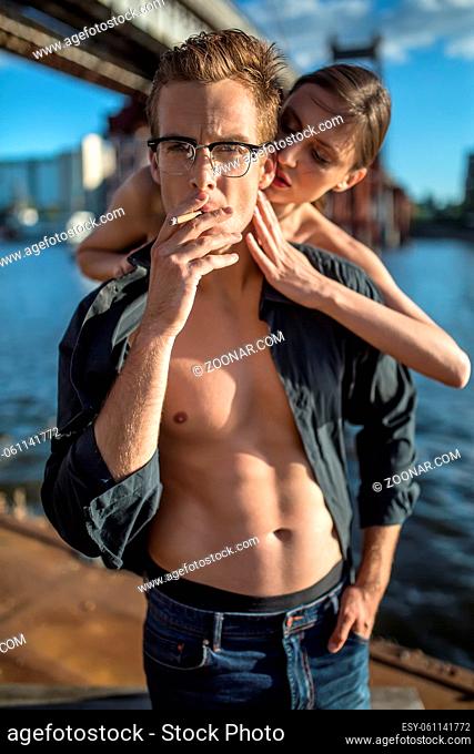 Trendy couple stands on the quay on the background of the bridge and the river. Girl hugs the guy from behind. Man wears a dark unbuttoned shirt