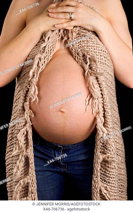 A pregnant woman wearing blue jeans clutches a brown blanket around her, exposing her bare belly. No face is shown