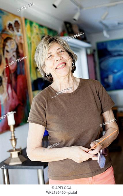 Smiling senior female artist holding paint brushes in studio