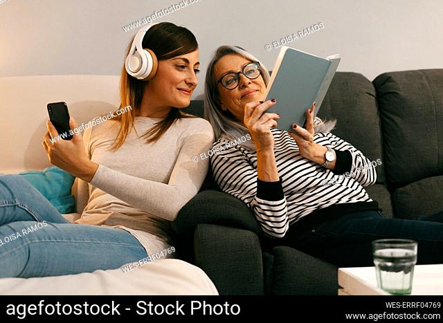 Mother showing book to daughter using mobile phone while sitting on sofa at home