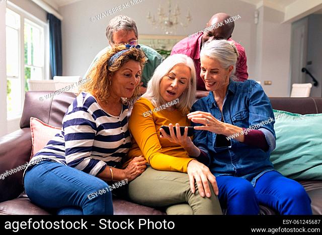 Surprised multiracial senior female friends looking at smart phone while men peeking from behind