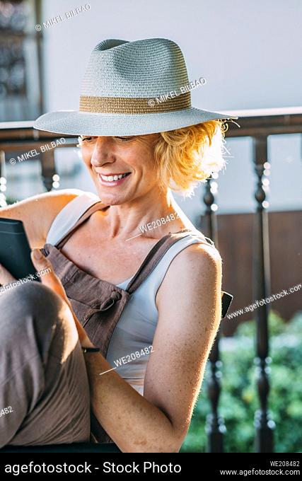 portrait of a mature caucasian blonde young woman in her 50s wearing a hat and reading an ebook and sitting in a rocking chair in a relaxed pose on the wooden...