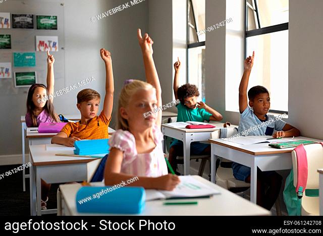 Multiracial elementary school students raising hands while sitting at desk in classroom