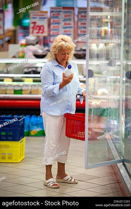 Senior woman shopping for food in the grocery store. She taking bottle of milk from the fridge and checking the label before putting it into the basket