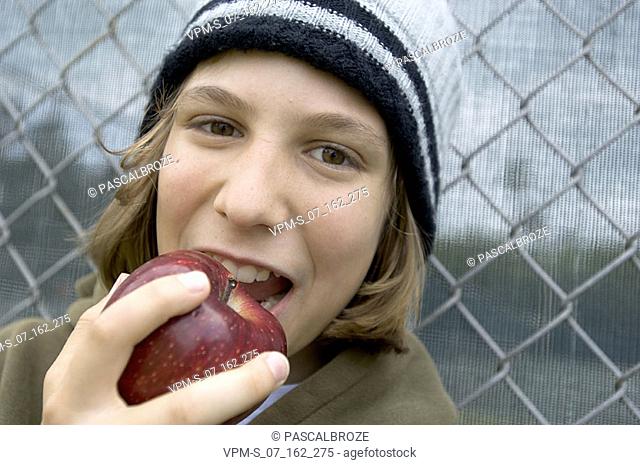 Portrait of a teenage girl leaning against a chain-link fence and eating an apple