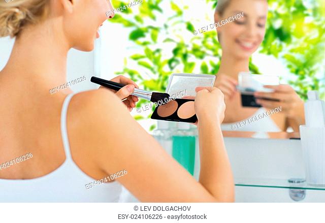 beauty, make up, cosmetics, morning and people concept - close up of happy young woman with makeup brush and contouring highlighting palette at bathroom over...