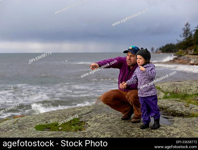 Father stands holding the hand of his young daughter as they stand at the water's edge looking out at the rough ocean water, Sunshine Coast, BC