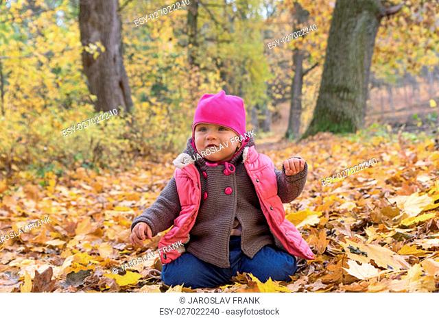 Baby girl in pink warm coat and pink hat is sitting on the ground in beautiful autumn park on fall day