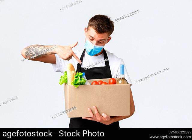 Covid-19, contactless shopping and groceries delivery concept. Handsome salesman in medical mask pointing at box with grocery order