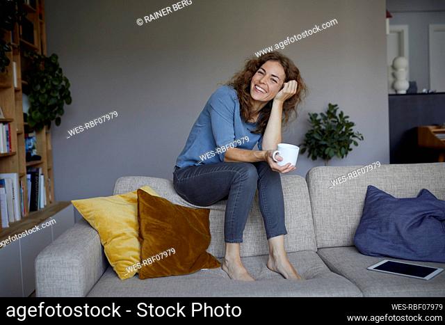 Smiling woman drinking coffee while sitting on top of sofa at home