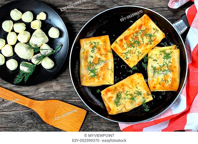 delicious mozzarella spinach stuffed crepes wraps on skillet sprinkled with finely chopped dill, on dark wooden board with mini mozzarella balls on black plate...