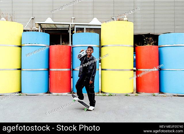 Happy young man with boom box and skateboard standing in front of concrete pipes