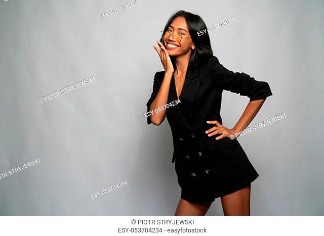 Portrait of attractive young Asian smiling woman in elegant black blazer and glasses posing over light gray wall background. Mock up copy space