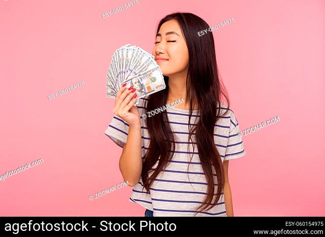 Portrait of attractive brunette girl greedy for money holding dollars and smelling banknotes with expression of much pleasure, enjoying wealthy life