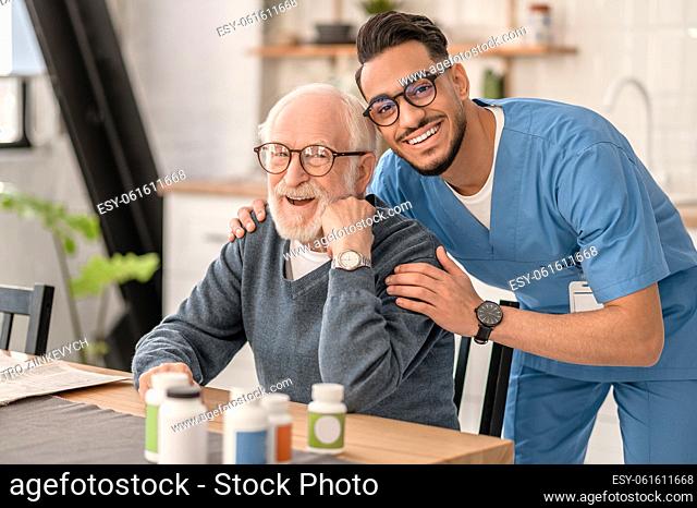 Smiling cheerful young medical worker dressed in uniform embracing a joyous elderly person from behind