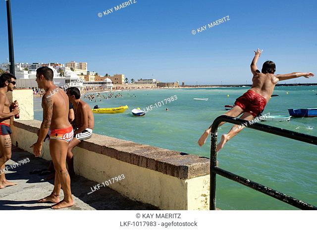 3 men on the beach and one boy jumping from the harbour wall into the sea next to the Castillo de Santa Catalina, Cadiz, Andalusia, Spain, Europe