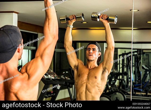 Handsome shirtless muscular young man exercising shoulders in gym with dumbbells