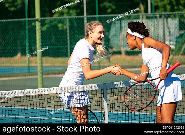 Happy biracial young female tennis competitors shaking hands over net at court on sunny day