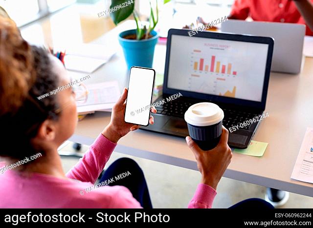 Businesswoman holding coffee cup while looking at smartphone with copy space in office