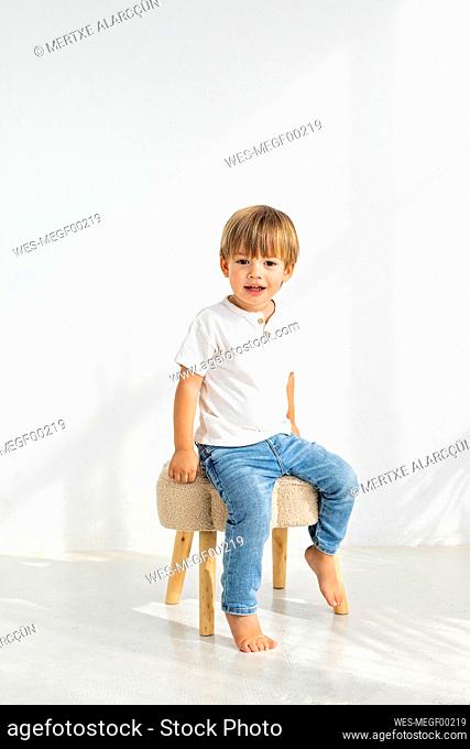 Cute boy sitting on stool in front of wall at home