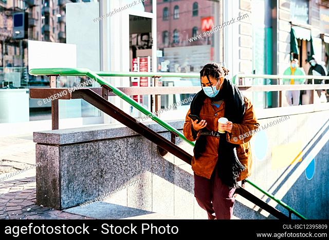 Italy, Woman in face mask holding smart phone and disposable cup on steps