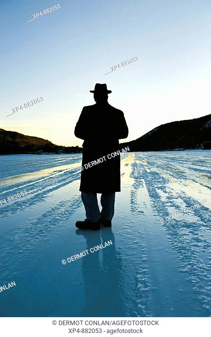 Silhouette of a man wearing an overcoat and a hat standing on a frozen lake strating off to the horizon, his back is to the camera