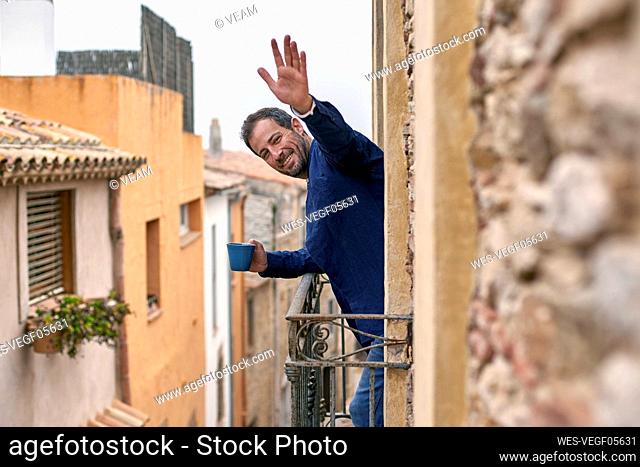 Happy man holding coffee cup waving from balcony