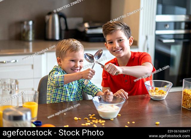 Smiling nice boy and his younger brother crossing their tablespoons over the bowl with cereal