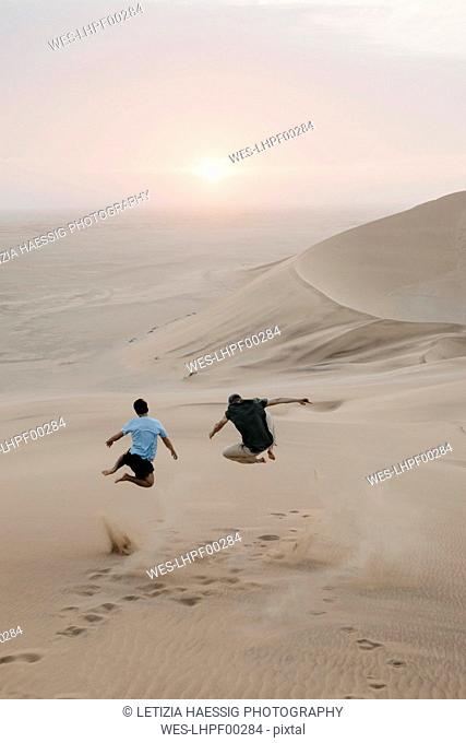 Namibia, Namib, back view of two friends jumping in the air on desert dune
