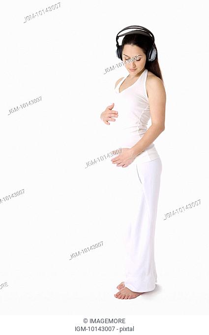 Pregnant young woman with headphones on, touching abdomen lightly, standing