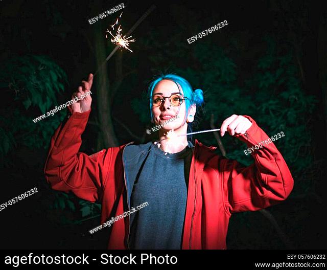 Portrait of hipster girl with blue dyed hair with sparklers in hands and reflection of it in glasses. Close up beautiful young teenage standing at night city