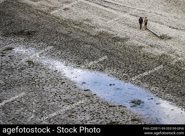 11 August 2022, France, Aiguines: Two men walk across the muddy shore at the Lac de Sainte-Croix reservoir during low water