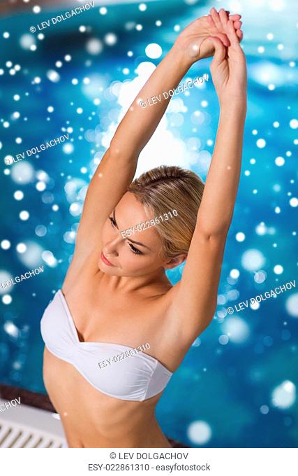 people, beauty, spa, healthy lifestyle and relaxation concept - beautiful young woman wearing bikini swimsuit sitting with raised hands in jacuzzi at poolside...