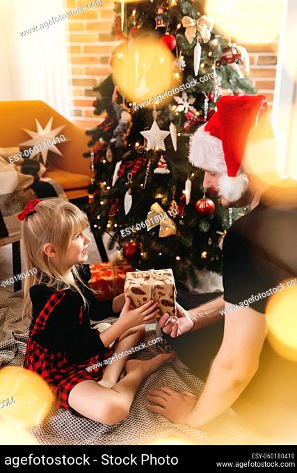 Young father in red Santa hat giving Christmas gift to his smiling daughter near decorated Christmas tree. Girl dressed in festive red-black Christmas outfit