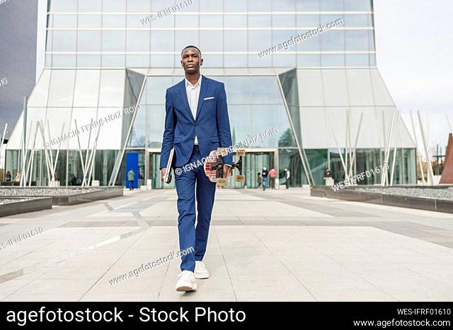 Young businessman holding skateboard and laptop walking in front of office building