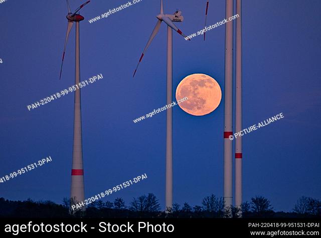 16 April 2022, Brandenburg, Petersdorf: The full moon shines at dusk between the masts of wind turbines in East Brandenburg. Photo: Patrick Pleul/dpa
