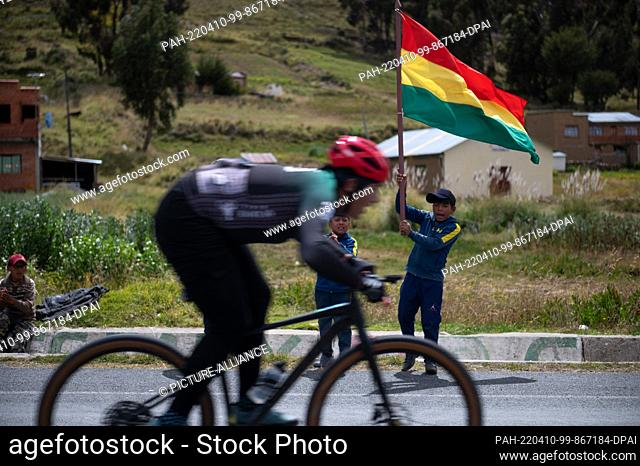 09 April 2022, Bolivia, Jankho Amaya: Children holding a Bolivian flag cheer on a cyclist during a bike race. The ""Poncho rojo"" (red poncho) race