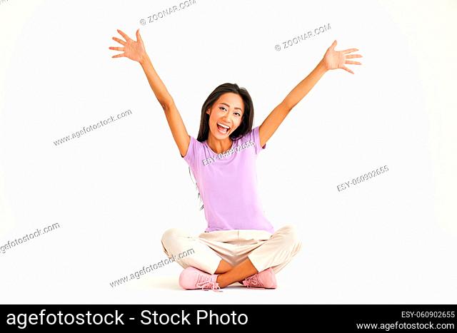 Smiling young asian woman raised arms up screaming and celebrating while sitting sitting on floor with legs crossed over white background