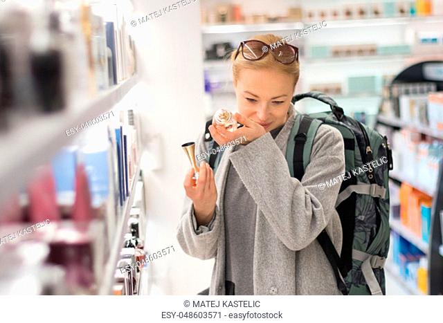 Blond young female traveler wearing coat and travel backpack choosing perfume in airport duty free store. Casual lady testing and buying cosmetics on the go in...