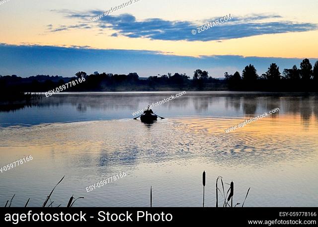 A fisherman in a boat floating on the water in the dawn sun in the fog