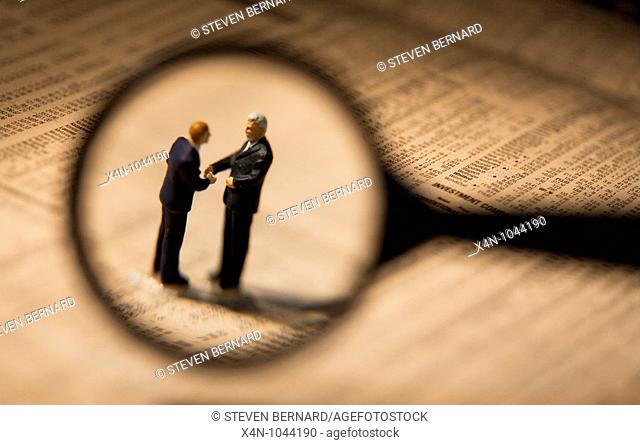 Miniature businessmen, seen through magnifying glass, shake hands while sstanding on financial newspaper