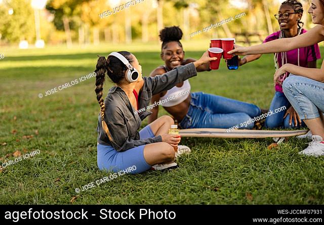 Young friends toasting disposable cups sitting at park