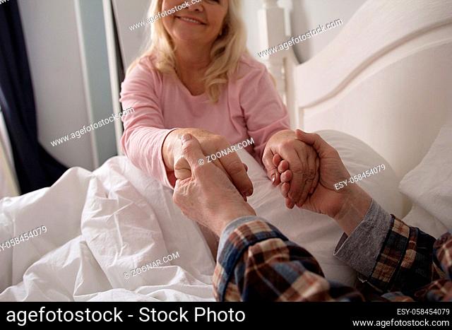 Cute old couple holding hands in their bed. Sweet old lady holding her husbands hands after waking up under white blanket