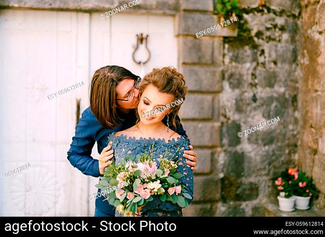 The groom kisses the bride on the cheek against the background of an old building with white doors in the old town of Perast . High quality photo