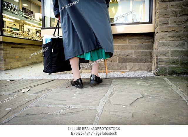 Back view of an unrecognizable old lady wearing a bag and a stick looking at shop window. Skipton, Yorkshire, England, UK